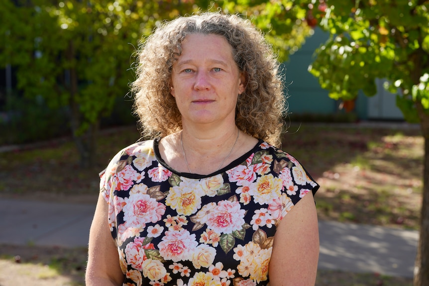 A woman with curly blonde hair in a floral print shirt stands outside looking serious.