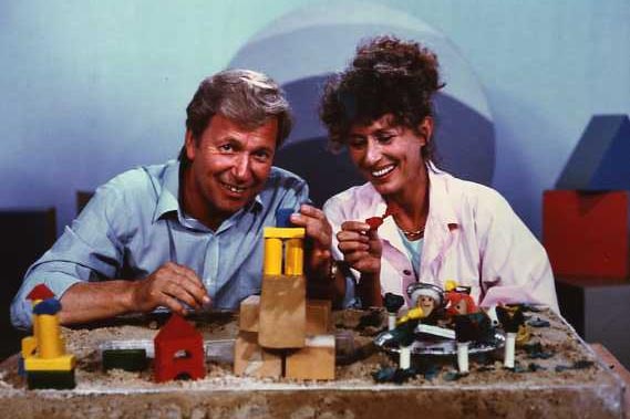 John Hamblin wears blue coloured shirt, smiling. Next to him Benita in pink coloured shirt, smiling. Wooden blocks on table.