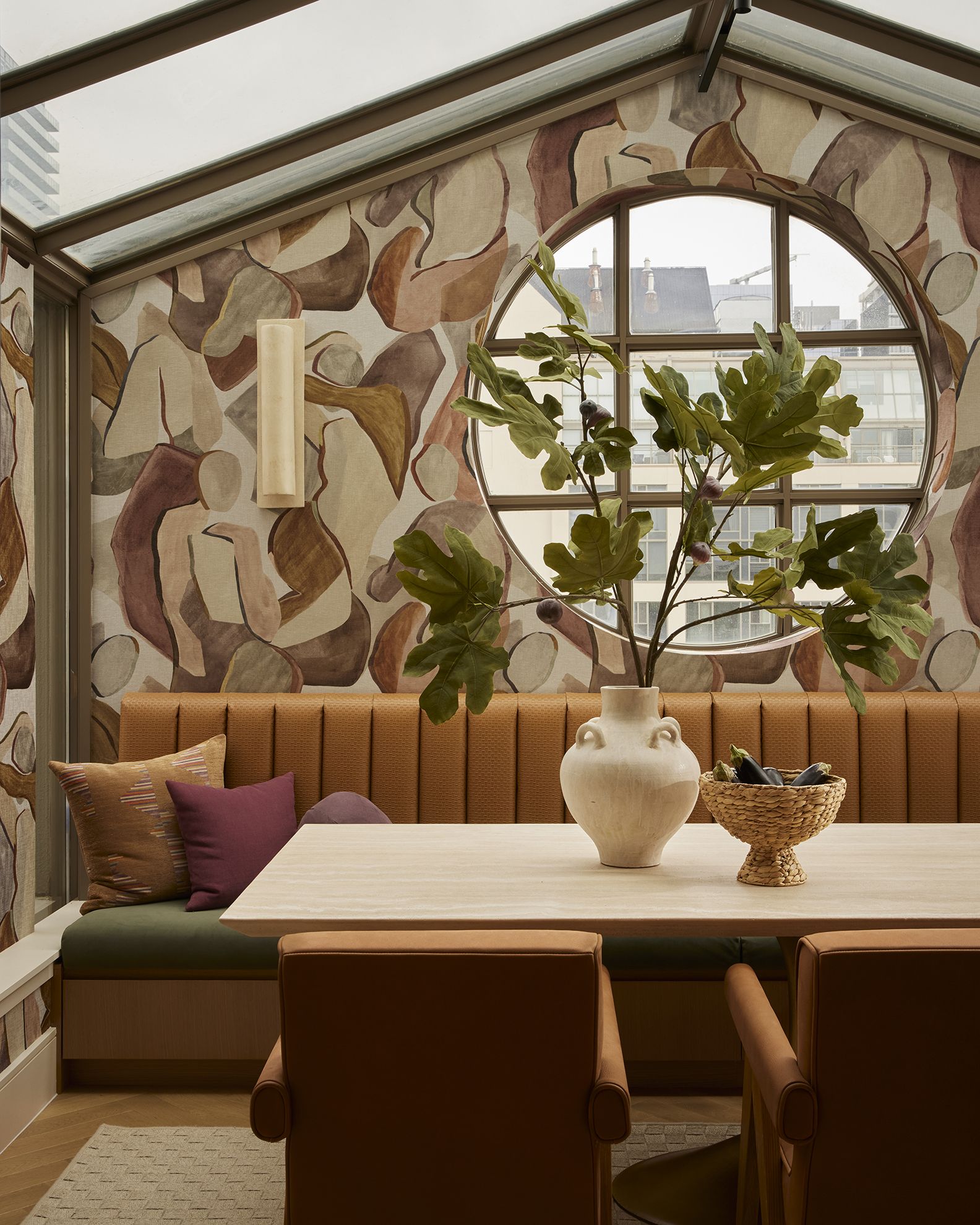 A veranda with a round porthole window, brown leather seating, mural-painted walls in tones of pink, brown, and beige, a central ceramic vase filled with fig leaves, and brown and bordeaux cushions.
