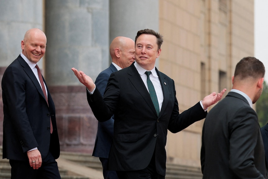Elon Musk wearing a suit holds his hands in the air on a red carpet looking happy.