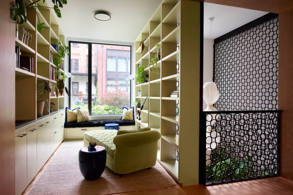 Modern living space with green chaise lounge, bookshelves, and large window showcasing outdoor greenery.