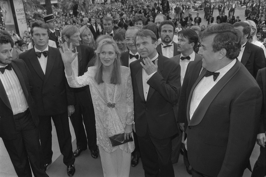A young Meryl Streep and Sam Neill in formal wear waving, surrounded by a group of suited men.