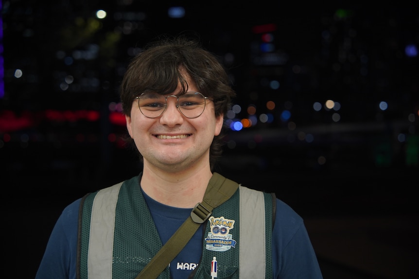 A man with glasses and dark hair smiles against a night time backdrop