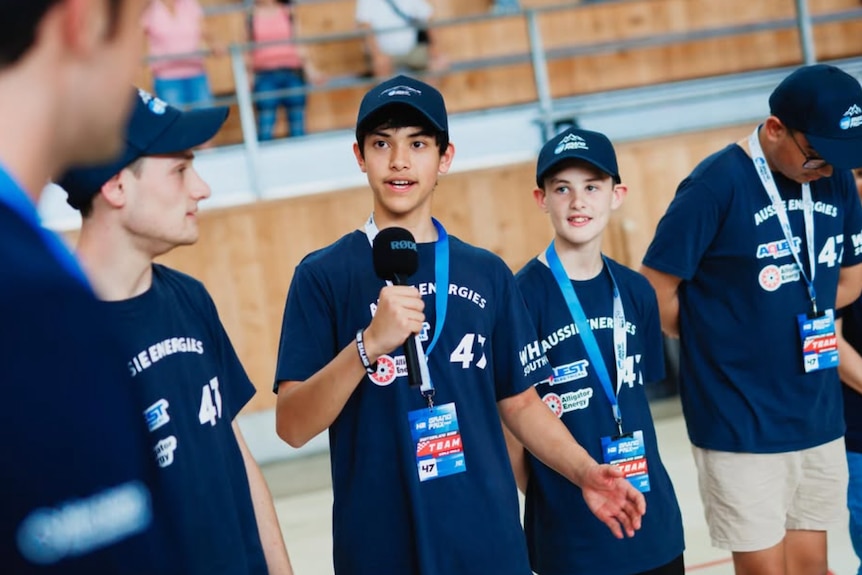 A teenage boy talking into a microphone for an interview flanked by his teammates