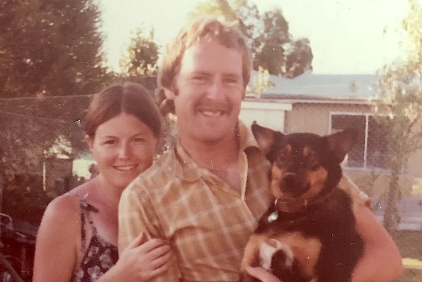 A young woman and man stand in a garden and smile. The man is holding a dog. 