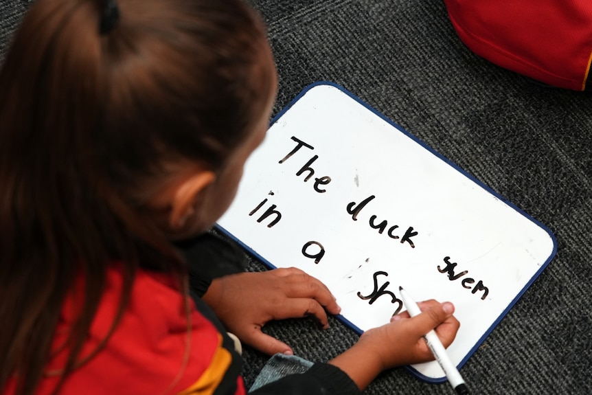 A school student with long hair in a pony tail, wearing school uniform leans over a white board holding a marker and writing.