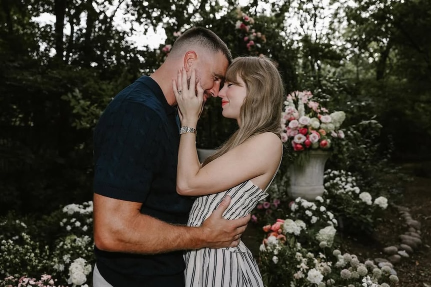 A man in a black shirt holds a woman wearing a black and white dress in a warm embrace in a flower-filled garden