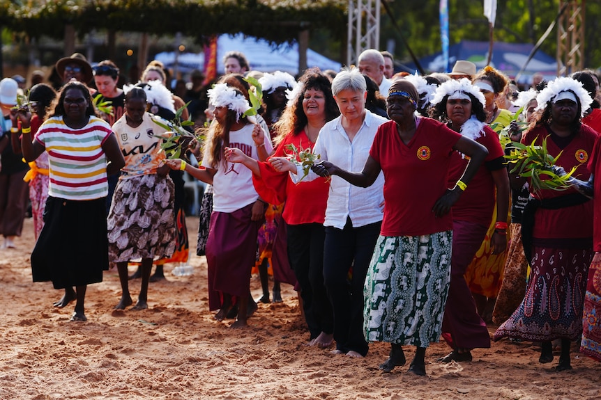 A woman with short, grey hair dances with Indigeous women in a bush setting.