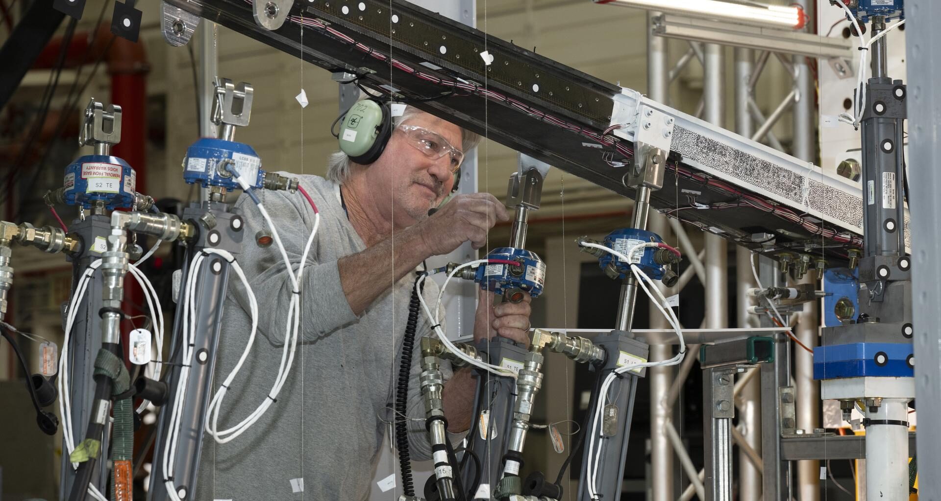 A man wearing ear protection works closely with multiple hydraulic and instrumentation units connected to a large beam mounted on a test structure. Numerous cables, hoses, and measurement devices extend from the setup.