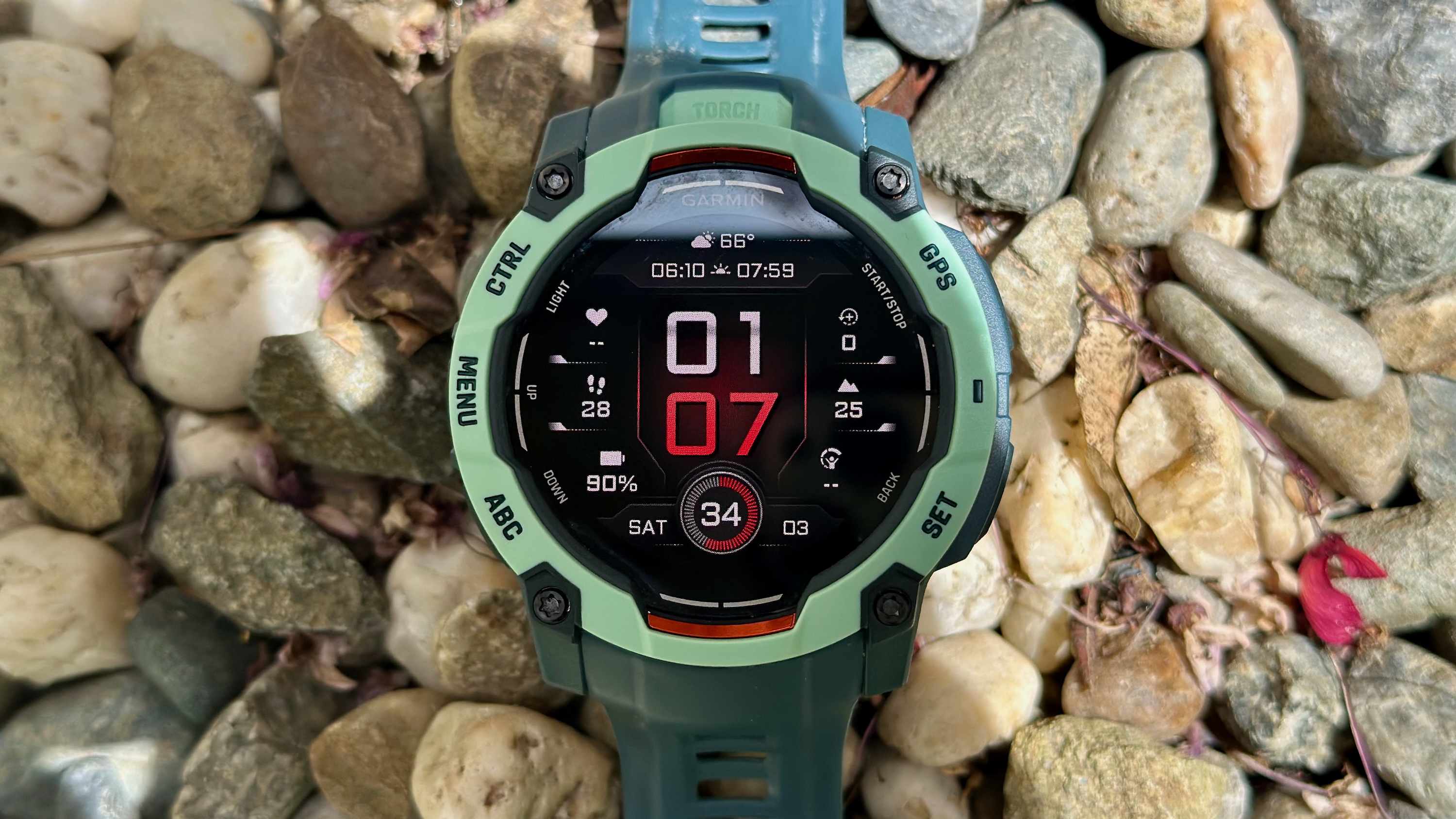 The Garmin Instinct 3 50mm AMOLED sitting atop pebbles and small rocks, the red-and-white watch face showing various data like the time &amp; date, weather, elevation, remaining battery life, and VO2 Max.