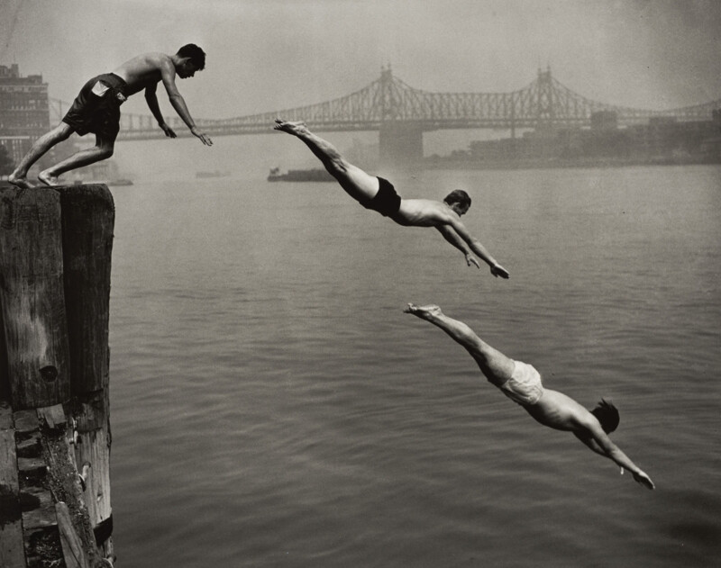 Three men dive into a river from a wooden pier, with a large bridge visible in the background.