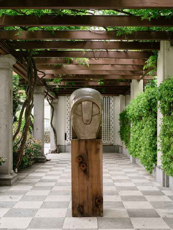 Stone sculpture on wooden base in covered outdoor walkway with checkered floor and green vine-covered pergola.