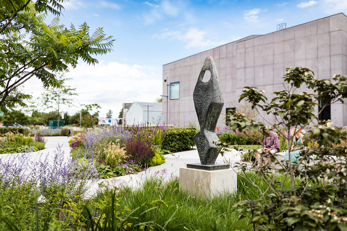 Modern sculpture in a landscaped garden with a concrete building in the background, surrounded by lush greenery and flowers.