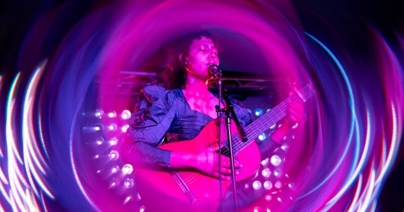 A musician plays guitar and sings into a microphone on stage, surrounded by vibrant pink and purple swirling light effects.