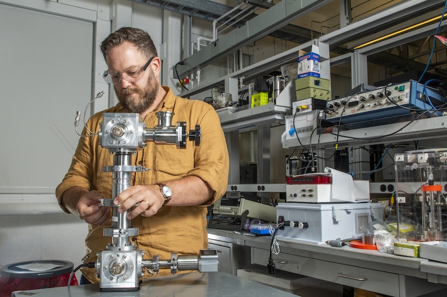 A man in a light brown shirt stands in a laboratory space in front of a metallic instrument about 15 inches tall made of small chambers attached with piping.