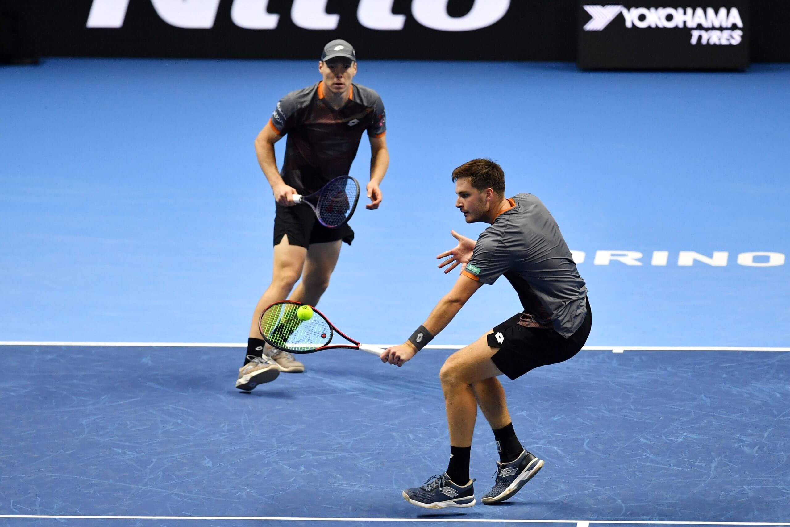 Harri Heliövaara (left) watches on as Henry Patten hits a left-handed backhand volley in front of him on a blue tennis court.