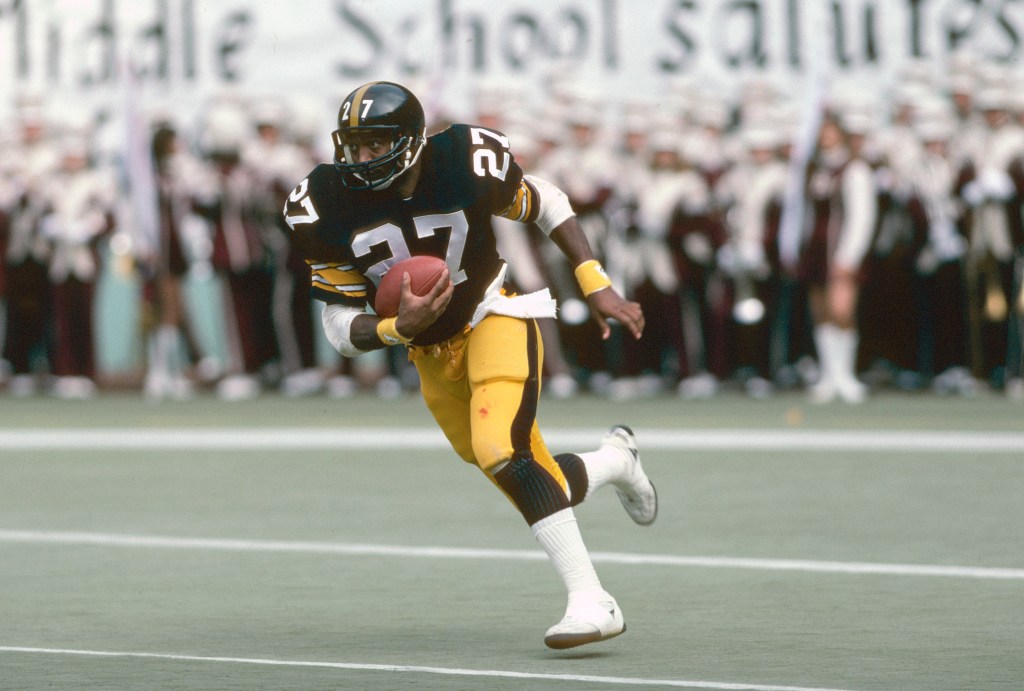 A football player in a black and gold uniform with the number 27, holding a football and running on a green field.
