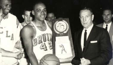 Walt Hazzard in a UCLA Bruins jersey holding a basketball, receiving a National Collegiate Basketball Championship trophy from John Wooden.