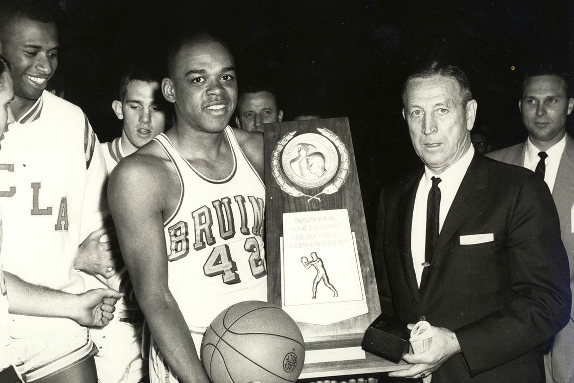 Walt Hazzard in a UCLA Bruins jersey holding a basketball, receiving a National Collegiate Basketball Championship trophy from John Wooden.