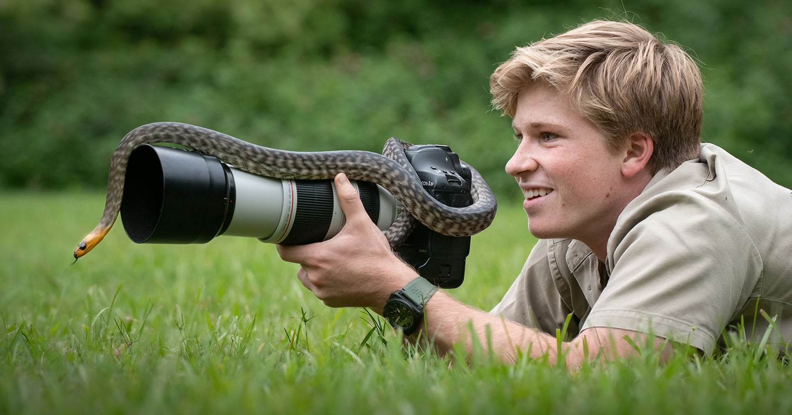 A smiling person lies on grass, holding a camera with a large lens while a snake is slithering over the lens; trees blur in the background.