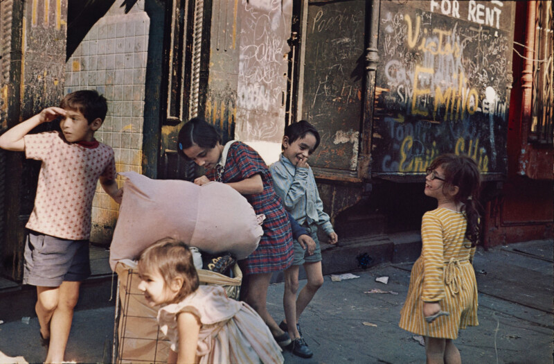Five children play on a city sidewalk in front of a building with graffiti and a "FOR RENT" sign.