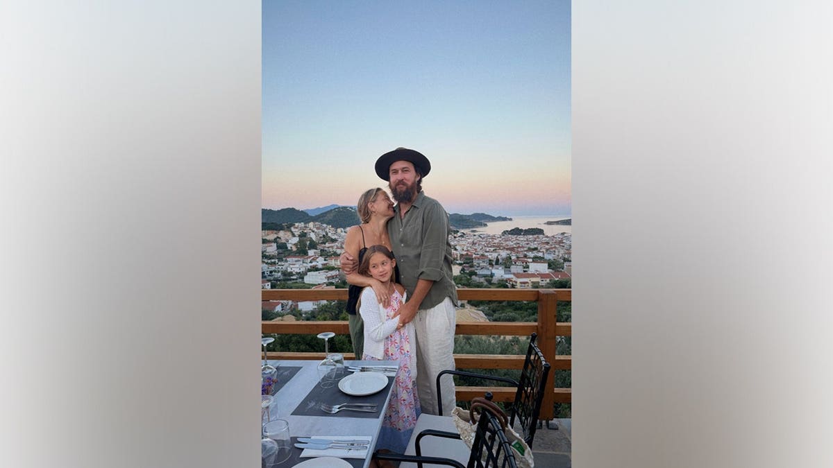 Kate Hudson smiles alongside fiancé Danny Fujikawa and daughter Rani during an outdoor dinner overlooking the water in Greece.