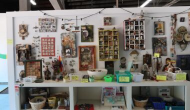 Shop display of handmade craft items: framed photos, jewelry, trinkets, and small boxes arranged on shelves with price tags and baskets beneath.