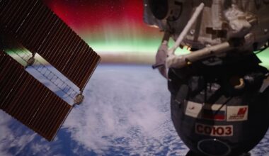 A view from the International Space Station shows a Russian Soyuz spacecraft and solar panels above Earth, with a glowing green and red aurora visible over the planet’s horizon.