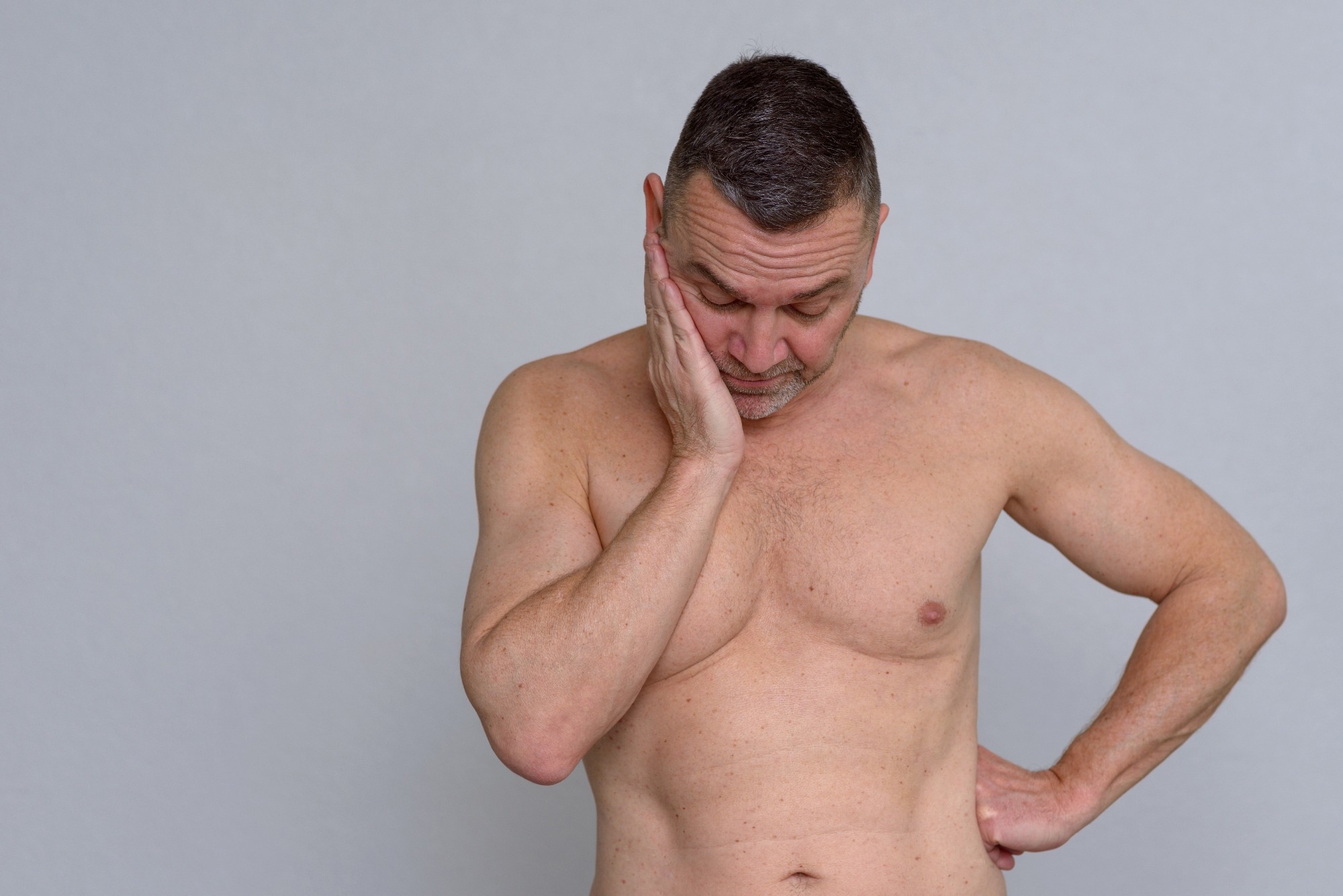 A studio portrait of a mature man naked from the waist up looking fed up and depressed.