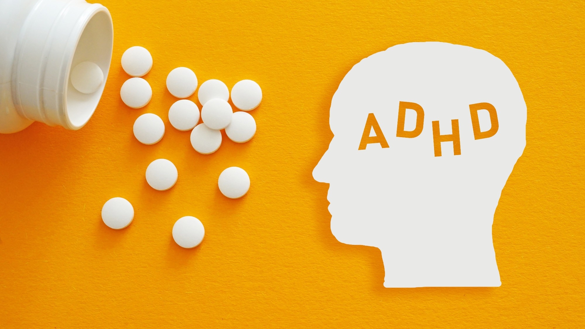 White ADHD head silhouette beside scattered medication tablets on a yellow background, illustrating stimulant treatment for attention deficit hyperactivity disorder.