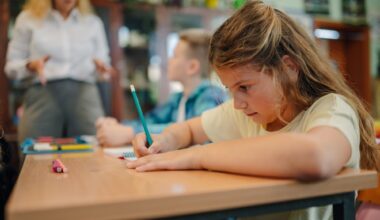 Schoolgirl writing on notebook with pencil during class, while teacher explains lesson to elementary school students