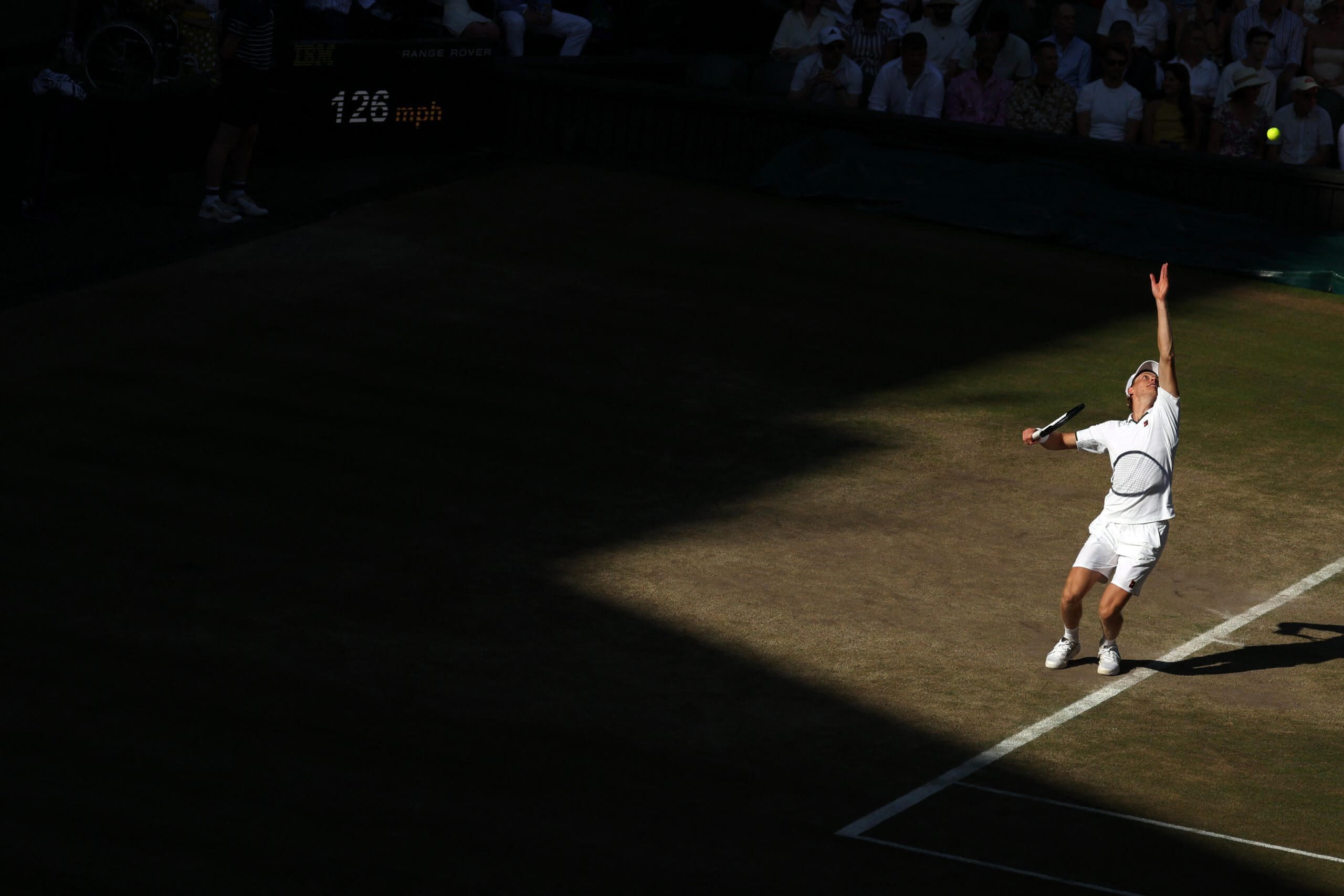 Jannik Sinner serves in a patch of light on a grass tennis court.