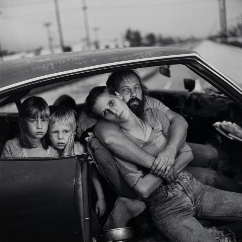 A black-and-white portrait of a family of four sitting inside a car, with a man embracing a woman while two young children look out from the back seat.