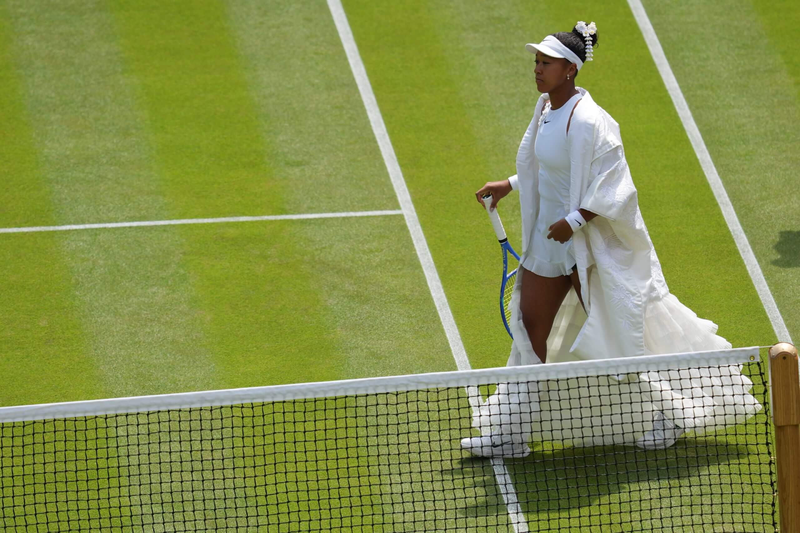 Naomi Osaka walks on to a grass tennis court wearing a flowing, white, kimono-style dress.