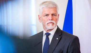 A man with white hair and a beard wearing a gray suit, white shirt, and blue striped tie stands in front of a blue and white flag, looking serious.