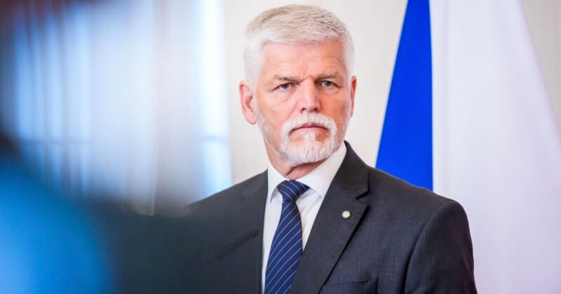 A man with white hair and a beard wearing a gray suit, white shirt, and blue striped tie stands in front of a blue and white flag, looking serious.