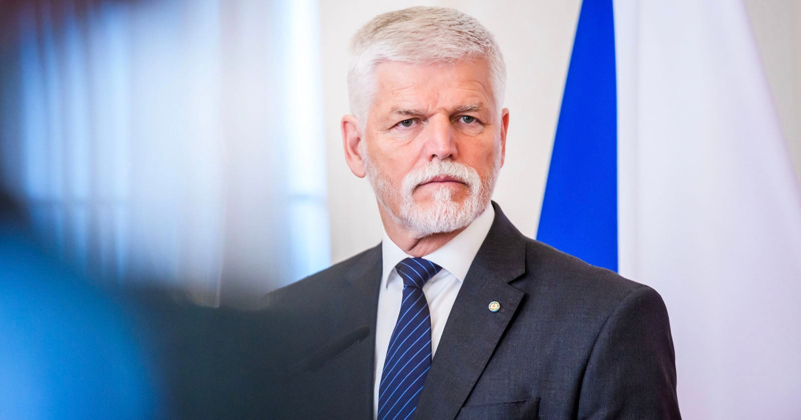 A man with white hair and a beard wearing a gray suit, white shirt, and blue striped tie stands in front of a blue and white flag, looking serious.