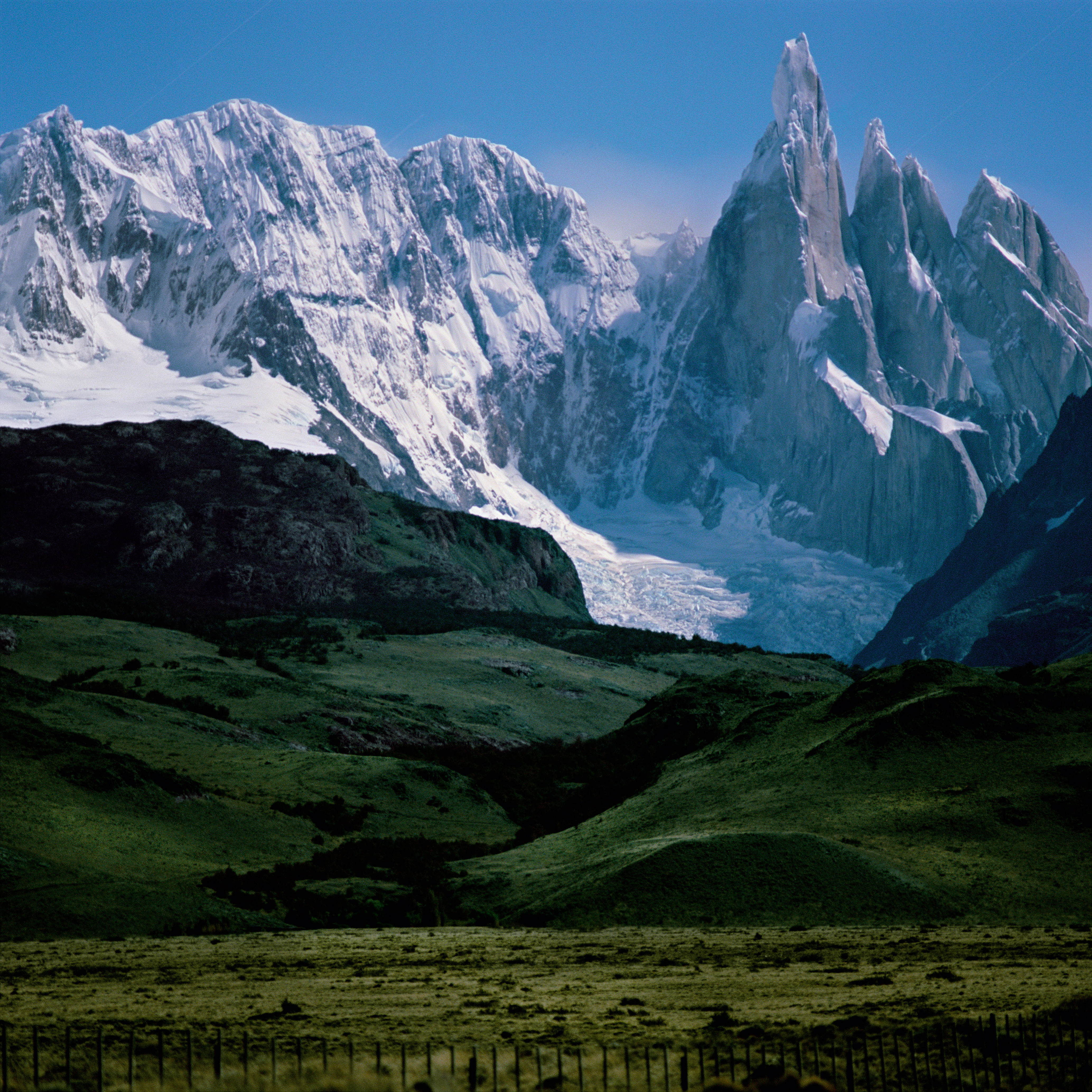 A striking mountain vista featuring jagged, snow-dusted peaks and a glacier rising sharply above lush green hills and a grassy plain.