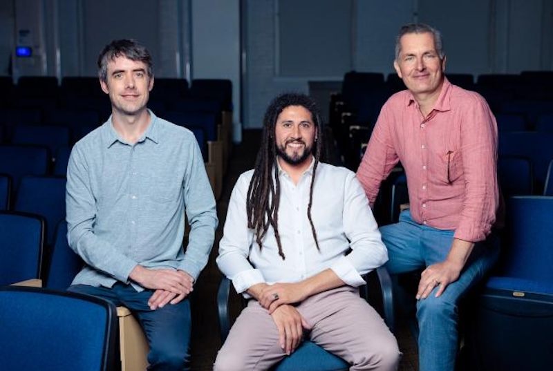 3 men sitting in blue seats. The middle man has long, braided hair.