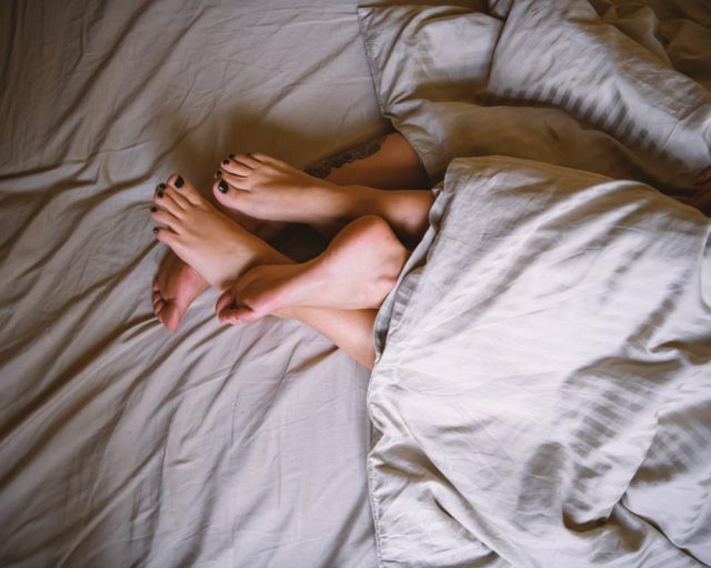 Lesbian couple lies in bed together, covered with a duvet while their legs are entwined
