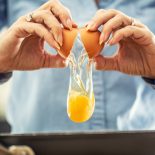 A detail of cracked egg falling into the pan as woman holds egg shells in both hands.