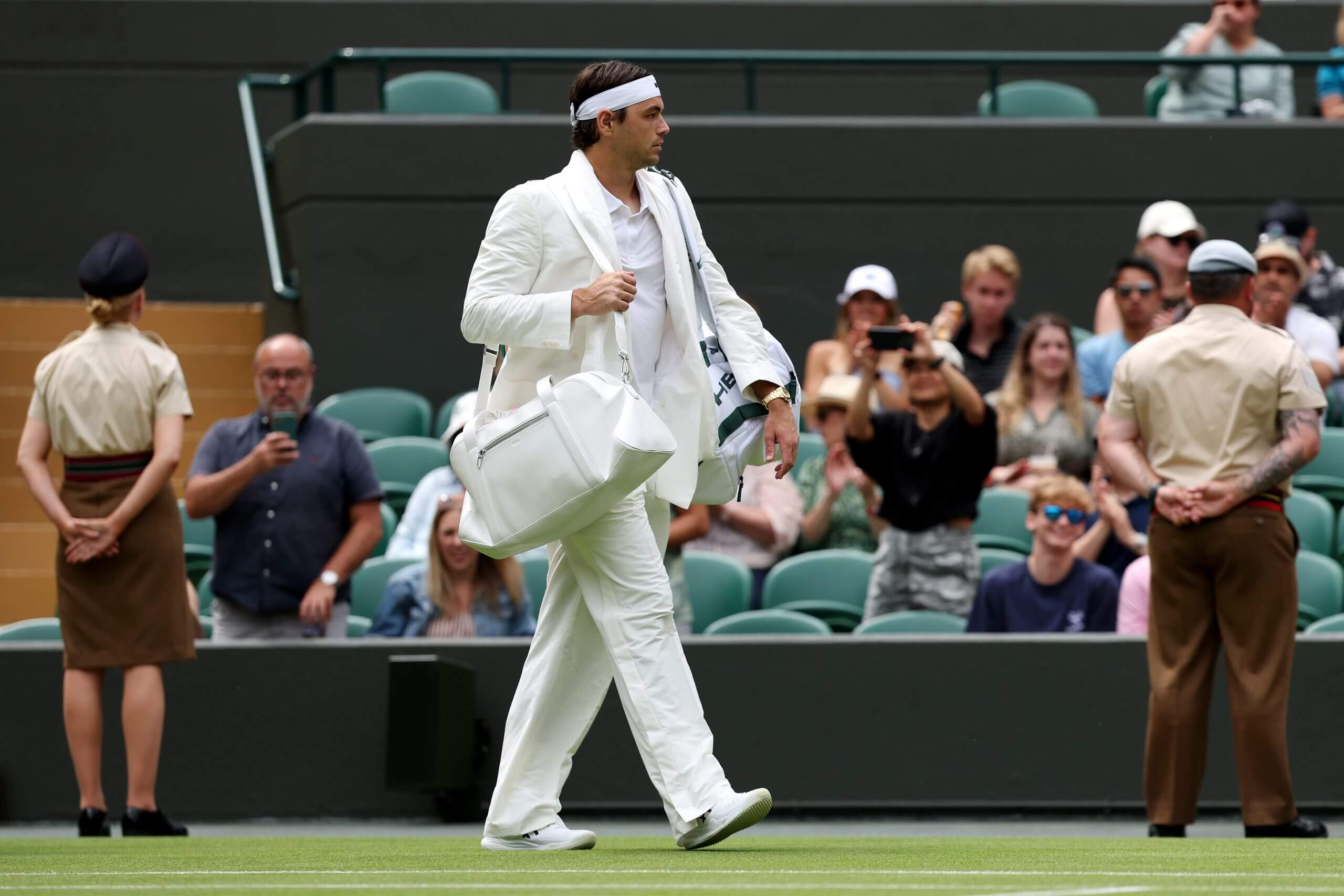 Taylor Fritz walks on to a grass tennis court wearing a white double-breasted suit and silk scarf.
