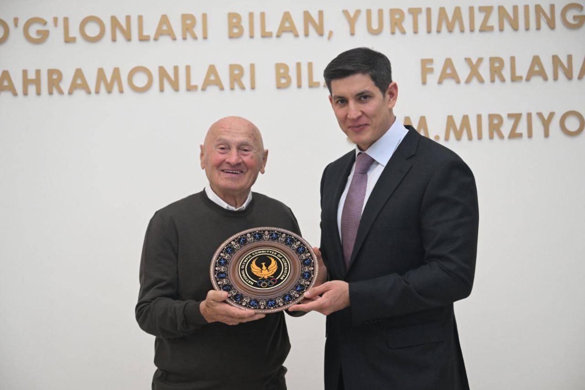 Reverberi, in a charcoal sweater, poses in front of a wall with Umarov in a dark suit. They are smiling and holding a commemorative plate in front of them.