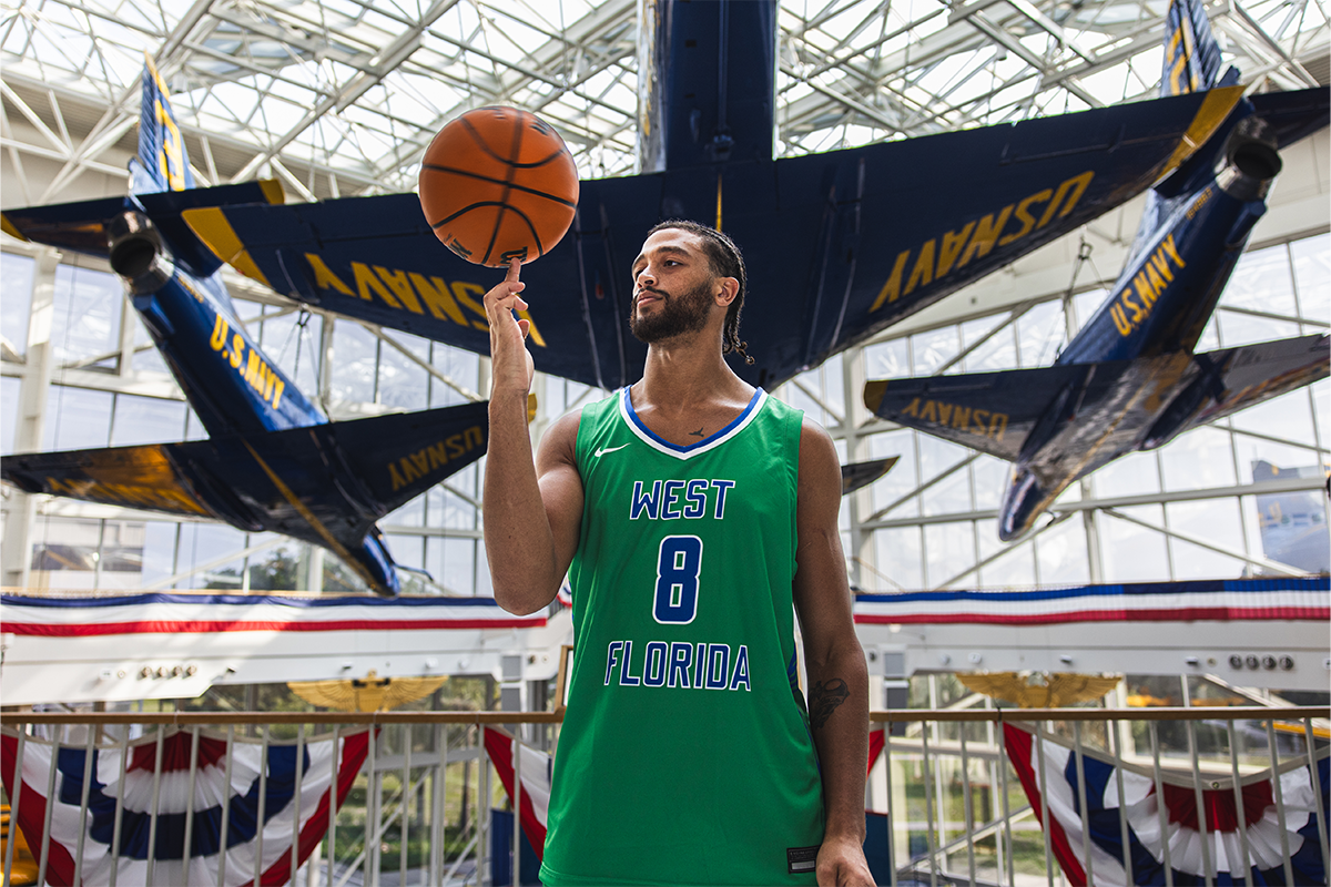 UWF men's basketball player with jersey number 8 spins a basketball on his finger while standing in front of a U.S. Navy Blue Angels aircraft.