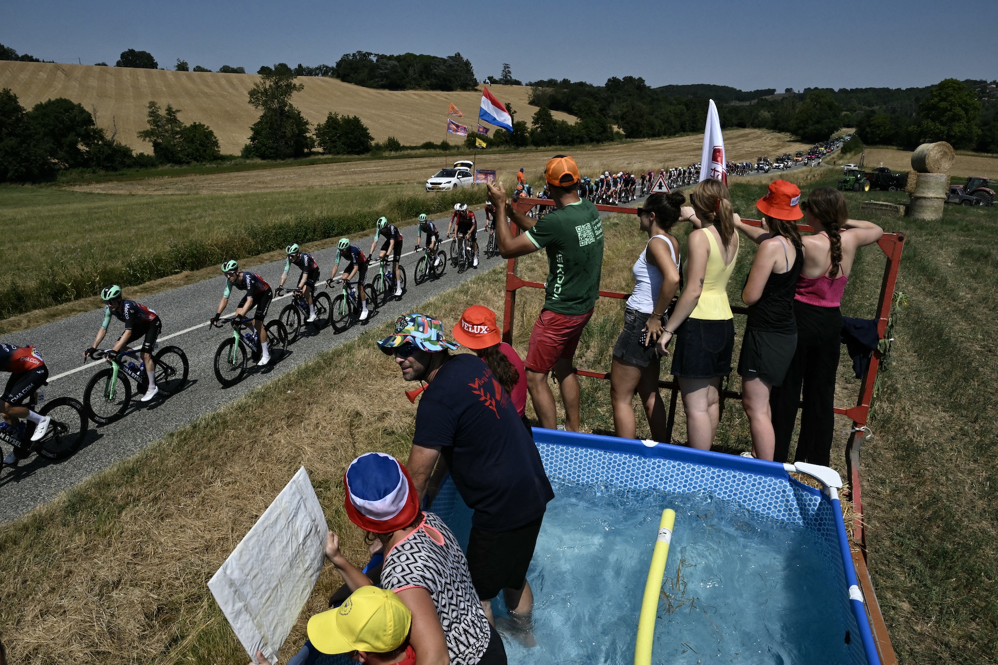 The pack of riders (peloton) cycles past spectators in a makeshift pool of water along the roadside during the 5th stage of the 113th edition of the Tour de France cycling race, 158,3 km between Lannemezan and Pau in southwestern France, on July 8, 2026. (Photo by Jeff PACHOUD / AFP)