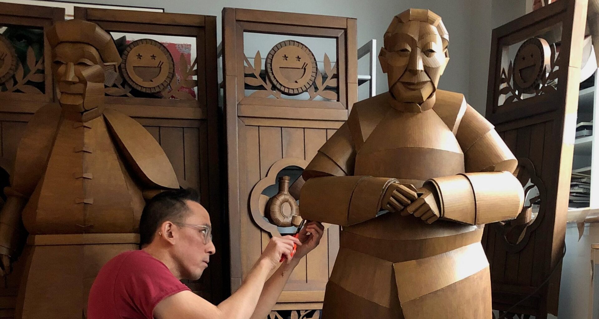 A person in a red shirt works on a large, brown, human-like sculpture in a room with wooden folding screens and another similar sculpture in the background.