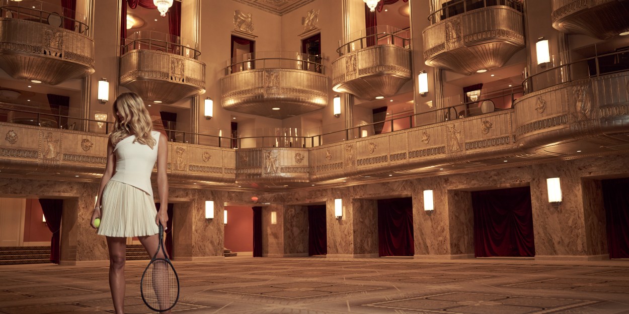 A lady in a white dress holds a tennis ball and racquet while in a grand ballroom