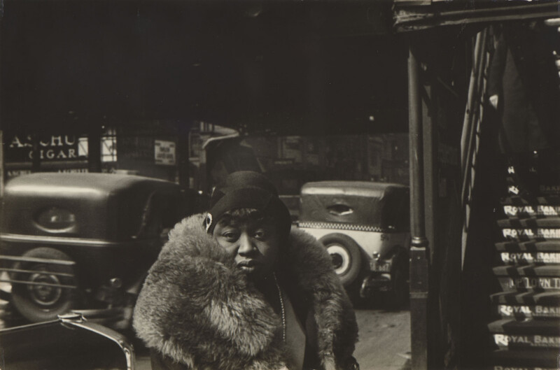 A woman wearing a dark cloche hat and a large fur collar stands on a city street with vintage cars and a staircase in the background.