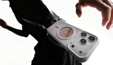 A person wearing black carries a white crossbody bag shaped like a smartphone with camera lenses and a small round screen displaying a cartoon face.
