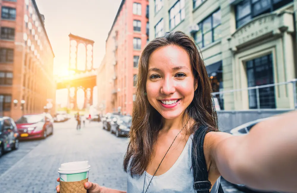 Gen Z female traveller holiding a takeaway coffee taking a selfie in NYC with Brooklyn Bridge in background.